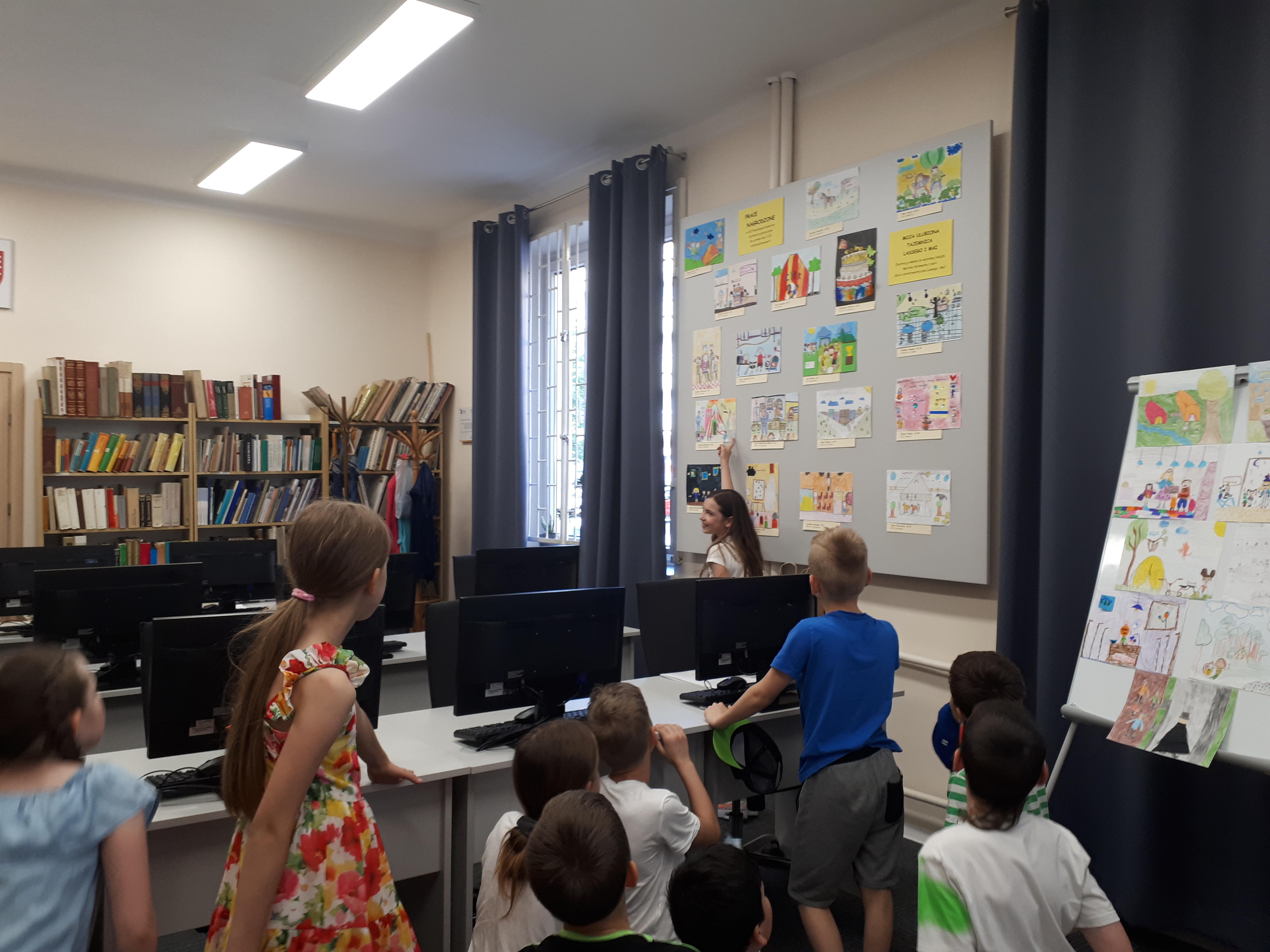 Sala biblioteczna z komputerami, dzieci oglądające wystawę prac nagrodzonych w konkursie.