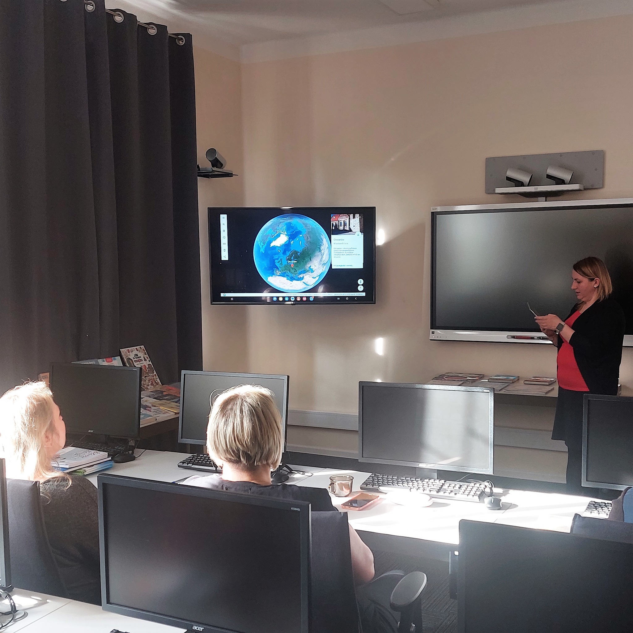 widok sali z osobami siedzącymi przy komputerach oglądających na ekranie telewizora aplikacje z rozszerzoną rzeczywistością