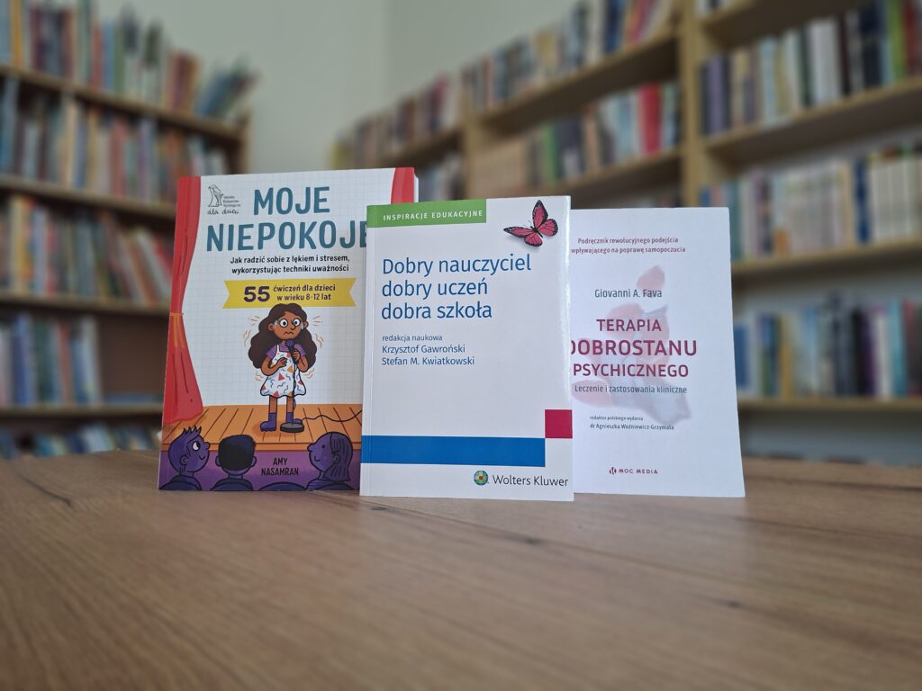 trzy książki ustawione na stole w tle zamazane regały z książkami